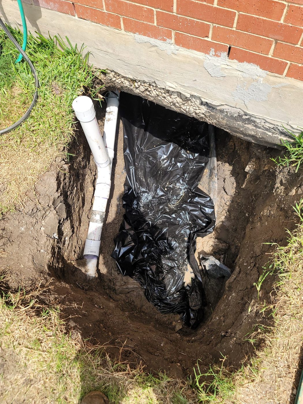 A tunnel dug at the exterior foundation of a brick house, with a new drain line and cleanout at grade